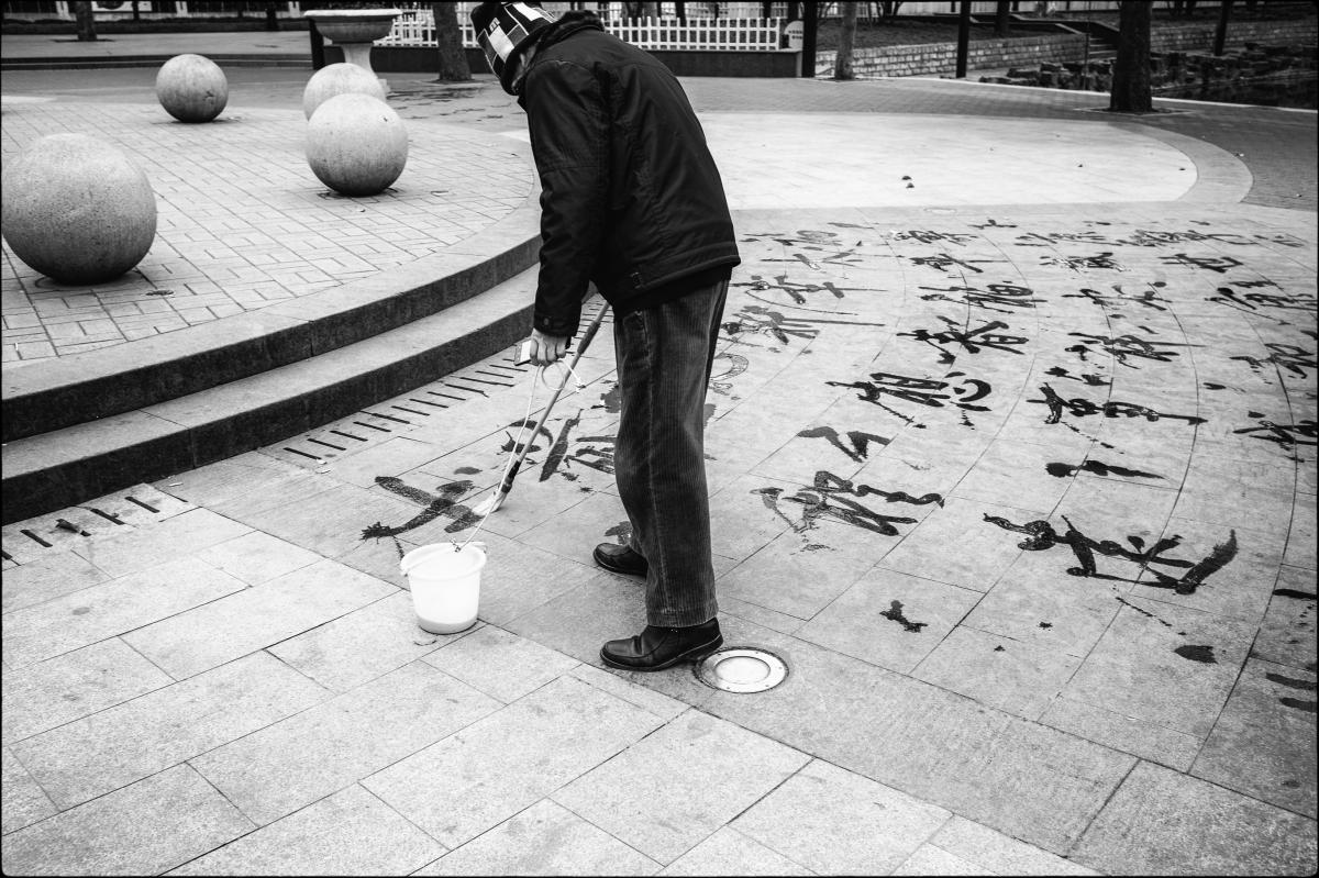 Without knowing what this man is doing, this is not a good photograph by any measure.  But when I learned what he was doing I had to try to capture it.  I did a poor job but here's the story.  I was working at a hospital in Suzhou, China, and was escorted everywhere by an orthopedic surgeon who went by the name of Edward.  Walking back to my hotel one afternoon I was intrigued by this man who carried a small pail of water and a long brush.  He was writing Chinese characters on the plaza of this park with water.  It was a cool day but the water soon evaporated, and then he started all over again.  I asked Edward what he was doing and he replied, "He just passing time."  That made no sense to me, so I watched him some more, despite Edward's imploring that we continue to our destination.  It then occurred to me that this man was practicing the Zen art of calligraphy.  With water.  That's like an entirely new level.  I said that to Edward who said he was unfamiliar with the term, Zen.  Just like another orthopedic surgeon told me she was unaware of anything that happened at Tiananmen Square.  I tried to explain it to him but he again said, "No, he just passing time."  Zen is not confined to the Seven Arts, is it?  Can photography be a Zen art?